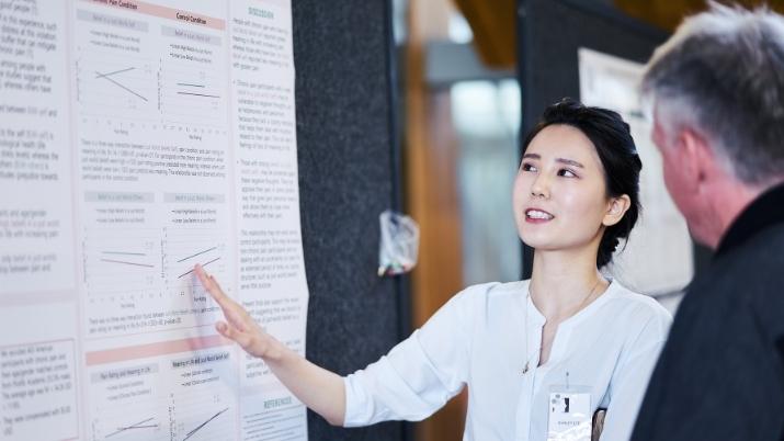 Picture with student standing in front of presentation board explaining her research to an onlooker. 