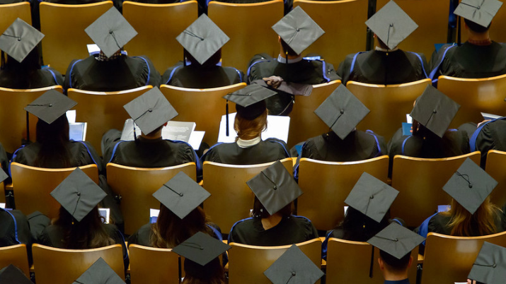 Image of students sitting in Chan Centre Hall, wearing black graduation gowns. The image is taken from a high point. 