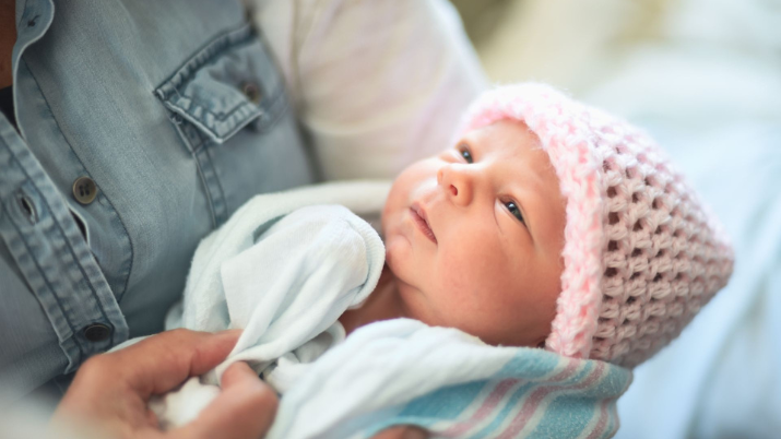  A newborn baby wearing a pink knit hat is swaddled in a hospital blanket and cradled in an adult's arms, gazing upward.